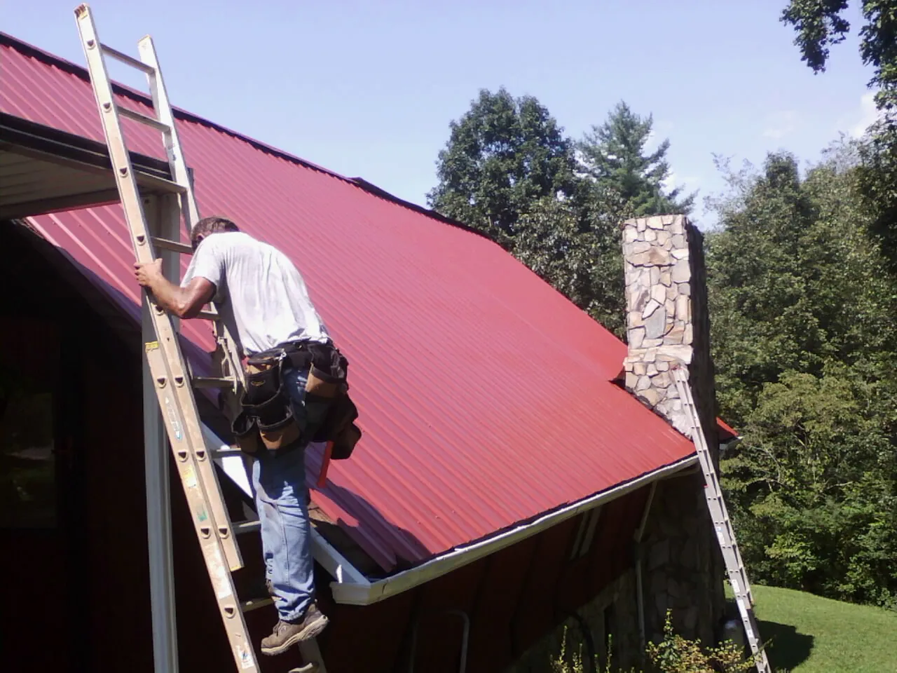 Roofer on a ladder beside a red metal roof and stone chimney — Soffit & Fascia Repair by Roofing Brothers in Taberg